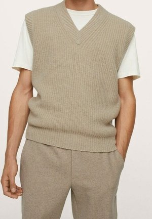 Beige gestrickte ärmellose Pulloverweste, getragen über einem weißen kurzärmeligen Hemd, kombiniert mit beigen maßgeschneiderten Hosen bei einer stehenden Person.