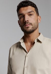 Beige, kort-gebouwde shirt met een knoop-kraag, met een subtiel logo op de borst. Gladde textuur; lichtgewicht stof.