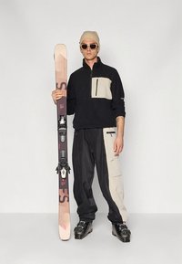 Sort og beige kvart-lynlåsfleece med en forlomme, beige og sorte skibukser, sorte skistøvler, holder lyserøde og brune ski.