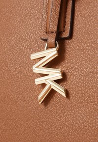 Sac en synthétique texturé marron avec un charme géométrique doré. Le charme présente un design triangulaire avec des angles vifs et une surface réfléchissante.