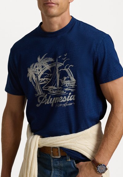 Marinblå bomullströja med ett silverfärgat grafiskt mönster av palmer, en segelbåt och texten "Polynesien." Bärs med en krämfärgad stickad tröja runt midjan.