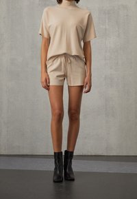 Haut en tricot beige à manches courtes, col montant et coupe ample, associé à un short à cordon de serrage assorti ; porté avec des bottines noires à la cheville.