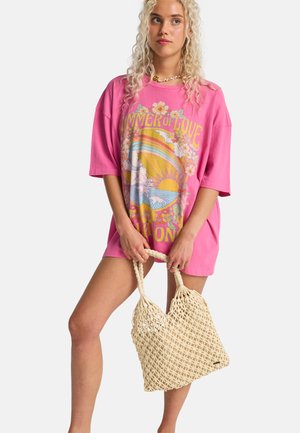 Jonge vrouw met krullend blond haar draagt een roze oversized T-shirt met een kleurrijke "Summer of Love" afbeelding, en houdt een beige netentas vast.