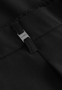 Zwarte stof met een gestructureerde uitstraling, met een zilveren metalen label gegraveerd met "GABBA" bevestigd aan de tailleband.