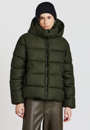 Grüne gesteppte Jacke mit hohem Kragen, Reißverschluss vorne und zwei seitlichen Taschen. Kombiniert mit einer schwarzen Strickmütze. Glatte Textur, verkürzte Passform.