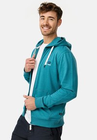Sweatshirt à capuche teal avec poches avant, cordons blancs et logo sur la poitrine gauche. Le tissu semble doux et légèrement texturé.