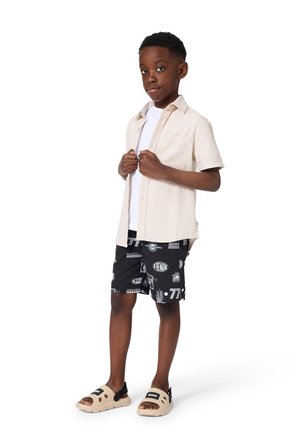 Junge trägt beiges kurzärmeliges Hemd über weißem T-Shirt, schwarze bedruckte Shorts und beige Sandalen, steht vor weißem Hintergrund.