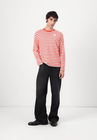 Rot-weißer gestreifter Langarmshirt mit Rundhalsausschnitt, kombiniert mit lockeren schwarzen Jeans und schwarzen Schuhen. Einfaches Logo-Detail auf dem Shirt.