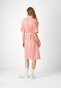Abito di lino rosa con maniche corte, vita cintata e orlo fino al ginocchio. Indossato con sneakers bianche e calzini alla caviglia. Vista posteriore.