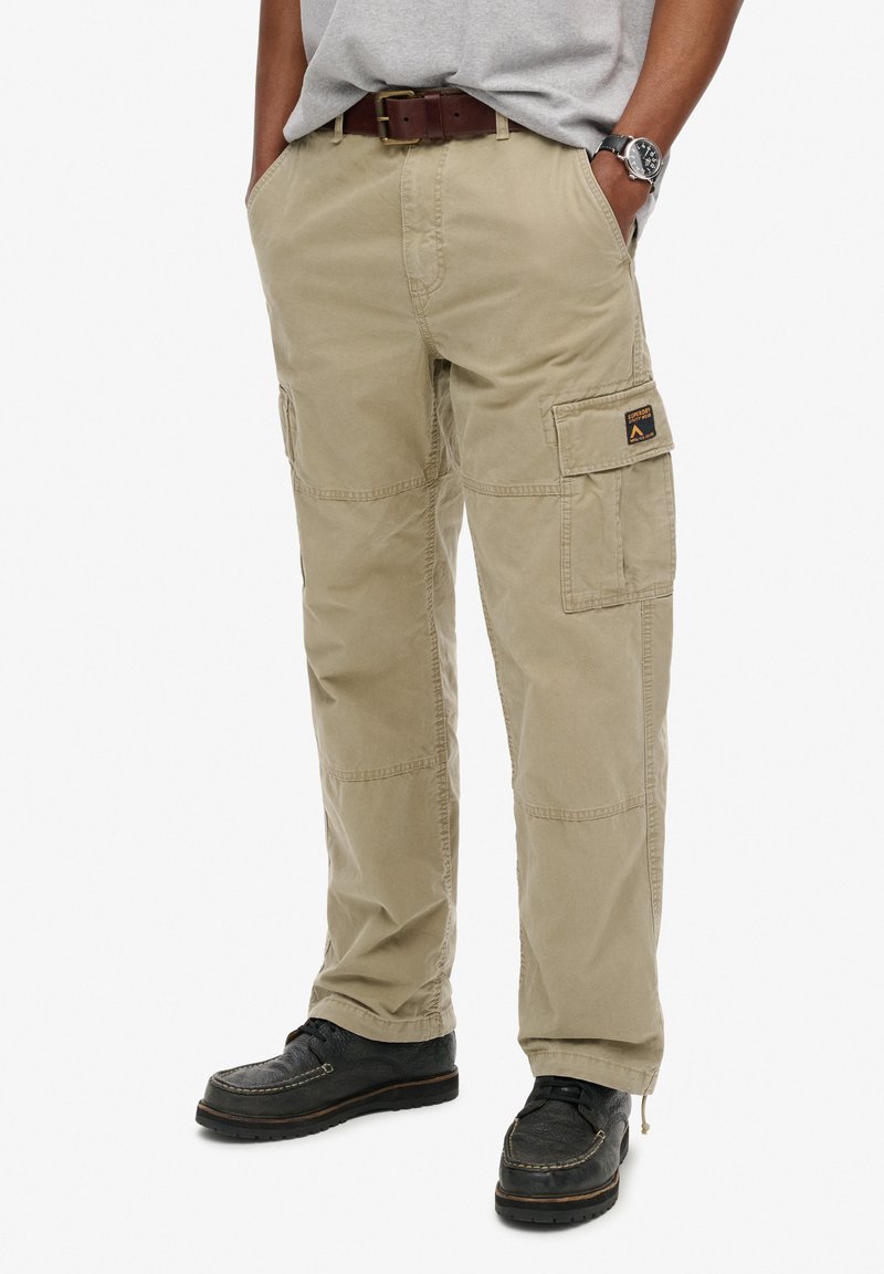 Beige cargo broek van duurzaam materiaal, met zijzakken en gestikte details, gedragen met zwarte schoenen. Eenvoudig, functioneel ontwerp.