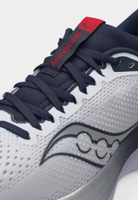 Κοντινή λήψη λευκού και ναυτικού αθλητικού παπουτσιού με ναυτικές κορδόνια και κόκκινο λογότυπο Saucony στην γλωττίδα, που δείχνει υφασμάτινη επιφάνεια με υφή και σχεδιασμό της μάρκας.