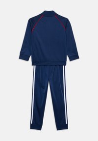 Marinblå träningsoverall med en zip-up jacka med röda detaljer och ribbade ärmslut, tillsammans med byxor som har vita sidostråk. Slät textur.