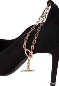 Chaussure à talon haut en daim noir, ornée d'une chaîne dorée avec un maillon circulaire et un fermoir toggle. La chaussure présente un bout pointu et un design élégant.