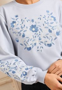 Svetlo modra trenerka z zapletenimi modrimi cvetličnimi vezeninami na sprednjem delu in rokavih, z rebrastimi manšetami in okroglo ovratnikom.