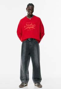 Rode gebreide trui met tekstontwerp, gecombineerd met losse donkere spijkerbroek. De look wordt compleet gemaakt met grijze sneakers met witte accenten.