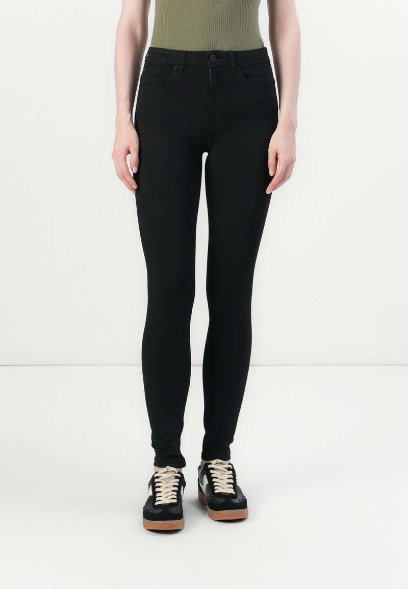 Schwarze enge Jeans mit glatter Textur, hoher Taille und Five-Pocket-Design, kombiniert mit schwarz-weißen Turnschuhen.