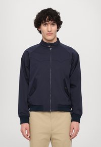 Giacca softshell blu navy con colletto alto, chiusura con zip, due tasche laterali, polsini a coste e una texture liscia. Design classico e funzionale.