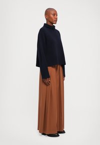 Kobieta stoi w brązowych szerokich spodniach, czarnych butach i luźnym granatowym swetrze z wysokim kołnierzem na jednolitym białym tle.