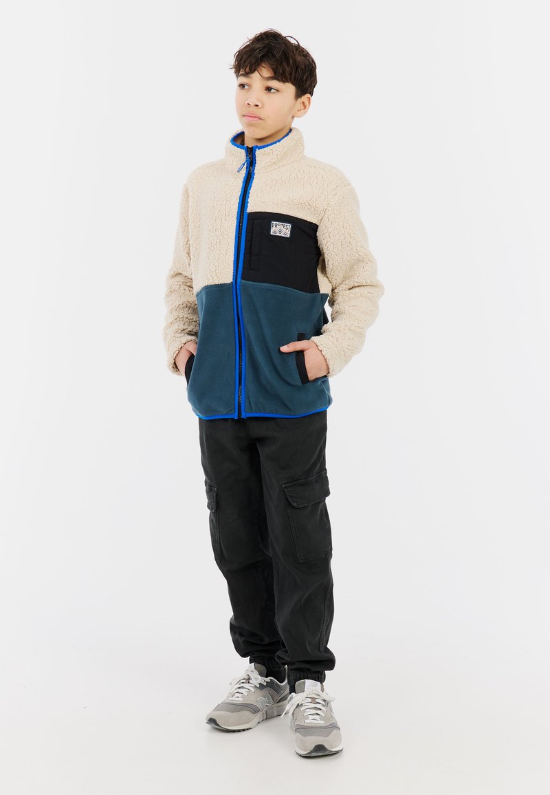 Fleece zip-up jacket in beige, black, and teal with a blue zipper, featuring a chest pocket and side pockets. Worn with black cargo pants and gray sneakers.