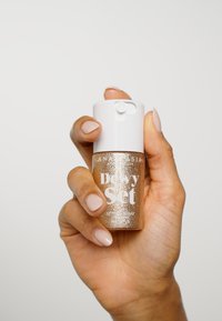 Spray utrwalający z brokatem w złotym kolorze w przezroczystej szklanej butelce z białą nakrętką. Etykieta mówi "Dewy Set" z tekstem w białym i złotym kolorze. Gładka tekstura.