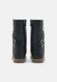 Bottines en cuir bleu marine avec une semelle en cuir tan, arborant une broderie de cœurs roses et un accent métallique. Fermeture à zip à l'arrière.