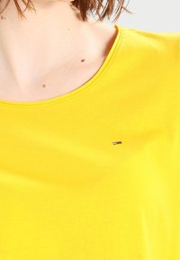 Žluté bavlněné tričko s kulatým výstřihem a malým značkovým logem v červené, bílé a modré barvě na hrudi. Hladká textura.