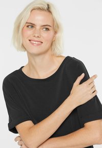 Young woman with short blonde hair wearing a black short-sleeve top, smiling and looking slightly to the side with arms crossed.