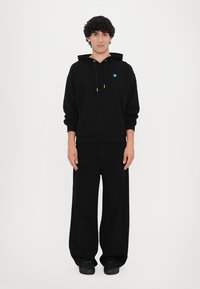 Felpa nera con cappuccio e tasca frontale, con un piccolo logo blu. Indossata con pantaloni neri larghi e affusolati e sneakers nere testurizzate.