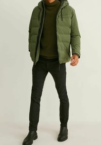 Doudoune verte avec capuche, dotée d'une fermeture éclair à l'avant et de deux poches sur la poitrine. Portée par-dessus un pull texturé vert foncé et un pantalon noir à motifs.