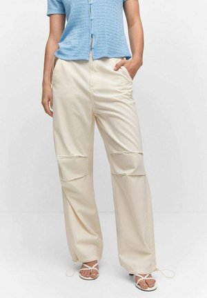 Persona con pantalones anchos color crema con parches en las rodillas, blusa de manga corta azul claro con botones y sandalias blancas de tiras, de pie con una mano en el bolsillo.