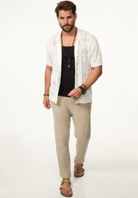 Weißes gehäkeltes Kurzarmshirt über einem schwarzen Tanktop, kombiniert mit beigen strukturierten Hosen und braunen Sandalen. Die Accessoires umfassen Halsketten und Armbänder.