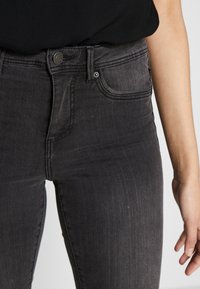 Vaqueros de denim negros de cintura alta con un ajuste slim, textura suave, bolsillos frontales y un detalle de cierre de botón. El acabado deslavado añade profundidad.