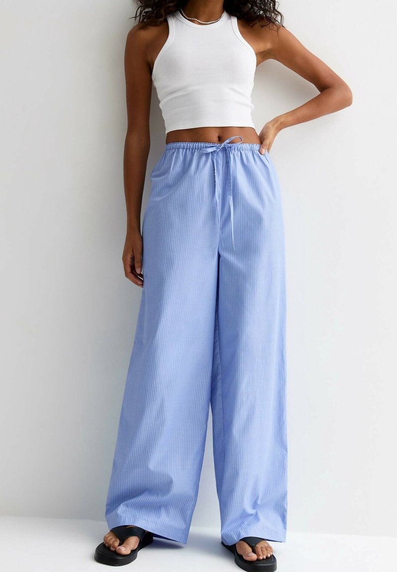 Vrouw in lichtblauwe wijde broek met een trekkoord in de taille, witte mouwloze crop top, zwarte sandalen en een parelketting.