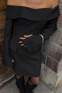 Črna off-shoulder obleka z dolgimi rokavi in širokimi manšetami, prilegajočim se pasom in gladkim materialom, v kombinaciji z črnimi škornji do kolen.