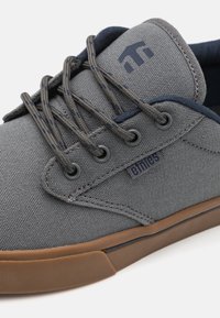 Sneaker din pânză gri cu accente bleumarin. Are șireturi rotunde, șase orificii pentru șireturi și o talpă din cauciuc. Un logo este vizibil pe lateral.