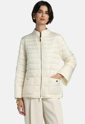 Kobieta ubrana w kremową pikowaną kurtkę z przednim zamkiem i kieszeniami naszywanymi, w połączeniu z pasującymi jasnymi spodniami i złotą biżuterią.