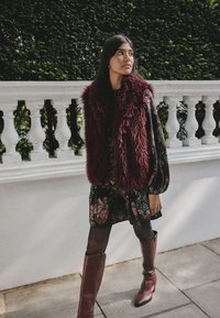 Burgunderfarbene Kunstpelzweste über einem geblümten Kleid mit langen Ärmeln, kombiniert mit burgunderfarbenen kniehohen Stiefeln auf einem steinernen Gehweg.