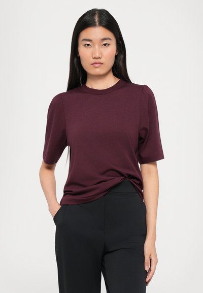 Woman with long dark hair wearing a dark burgundy short-sleeve top and black pants, standing with one hand in pocket against plain background.