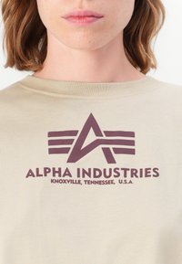 Beige bomull t-skjorte med en mørk lilla grafisk logo. Teksten inkluderer "ALPHA INDUSTRIES, KNOXVILLE, TENNESSEE, U.S.A." på forsiden.
