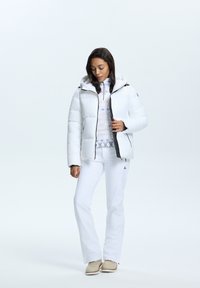 Doudoune blanche avec capuche, fermeture éclair et détails noirs. En dessous, un haut blanc à motif. Associé à un pantalon blanc et des chaussures beiges.