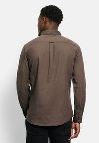 Mann mit kurzen, lockigen Haaren, der ein langärmliges braunes Hemd und schwarze Hosen trägt, von hinten vor einem schlichten weißen Hintergrund gezeigt.