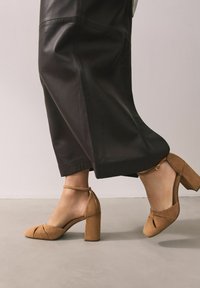 Hellbraune Wildleder-Riemchenpumps mit geschlossenem Zehenbereich, einem Riemen um den Knöchel und einem klobigen Absatz, kombiniert mit einem schwarzen Lederrock.