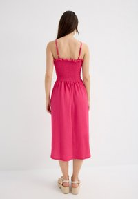 Vestido rosa de largo medio con un cuerpo fruncido, tirantes con volantes y una falda fluida. Combinado con sandalias de cuña visibles en la parte inferior.