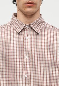 Κοντινό πλάνο ενός ατόμου που φοράει μπεζ και κόκκινο καρό πουκάμισο με κουμπιά και μυτερό γιακά, εμφανίζοντας μόνο τα χείλη και τον λαιμό.