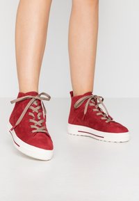 Czerwone zamszowe sneakersy za kostkę z beżowymi sznurowadłami, białą gumową podeszwą i czerwonymi akcentami. Charakteryzują się teksturowanym wykończeniem i sznurowanym fasonem.