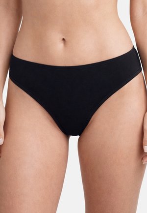 Mutandine nere a vita alta in stile bikini indossate sui fianchi di una donna, che mostrano l'addome inferiore e le cosce superiori su sfondo bianco.