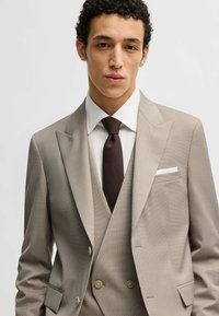 Completo beige su misura con finitura texture, gilet doppiopetto, camicia bianca e cravatta marrone. Presenta rever a punta e fazzoletto da taschino.