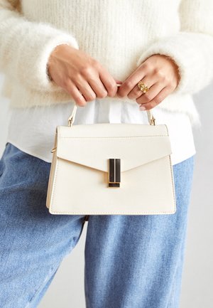 Krēmkrāsas rokassoma ar strukturētu formu, teksturētu materiālu, zelta detaļām un melnu akcentu uz aizdares. Regulējams siksniņa.