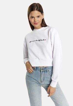 Witte cropped sweatshirt met een ronde hals, versierd met zwarte en roze borduring die "geef me een mooie glimlach" leest. Gestyld met blauwe jeans.