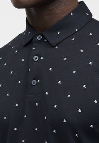 Κοντινό πλάνο σκούρου πόλο πουκαμίσου με μικρά λευκά επαναλαμβανόμενα λογότυπα "AX", τρία κουμπιά και μυτερό γιακά, που φοριέται από ένα άτομο.